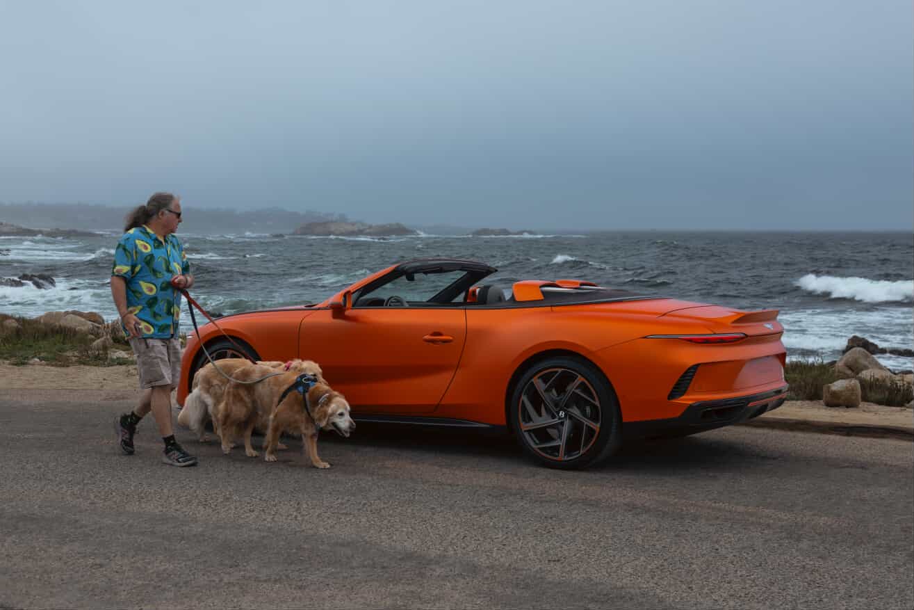 A picture of a man walking a dog past a sports car parked in front of the ocean.